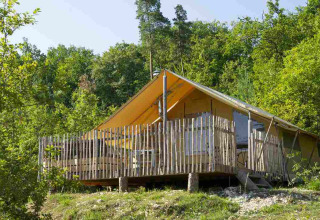 Safari tent with wooden deck and fence, nestled among green trees in a peaceful, forested landscape.