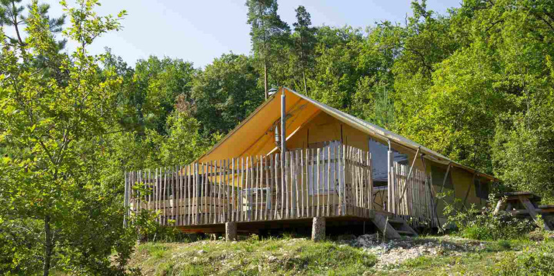 Tienda de safari con terraza de madera y cerca, ubicada entre árboles verdes en un entorno natural.