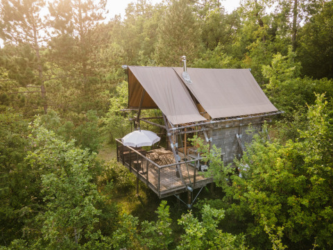 A Cahutte safari tent nestled in lush woodland, elevated with a deck, umbrella, and chairs among the trees.