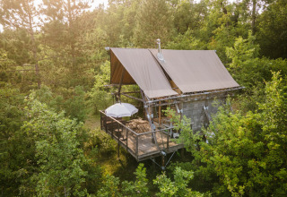 A Cahutte safari tent nestled in lush woodland, elevated with a deck, umbrella, and chairs among the trees.