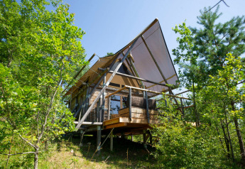 Safari teltet Cahutte i skoven, hævet på pæle med overdækket terrasse og omgivet af frodige grønne træer.