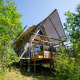 Tienda safari Cahutte en el bosque, elevada sobre pilotes con terraza cubierta y rodeada de árboles verdes.