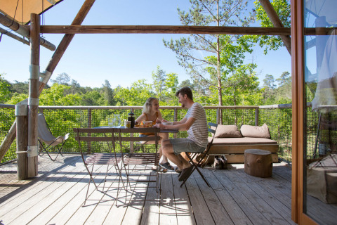 Couple enjoys a meal on the wooden terrace of a Cahutte safari tent surrounded by trees and nature.
