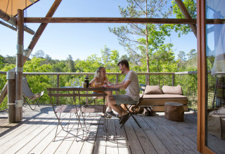 Pareja disfruta de una comida en la terraza de madera de una tienda Cahutte, rodeada de árboles y naturaleza.
