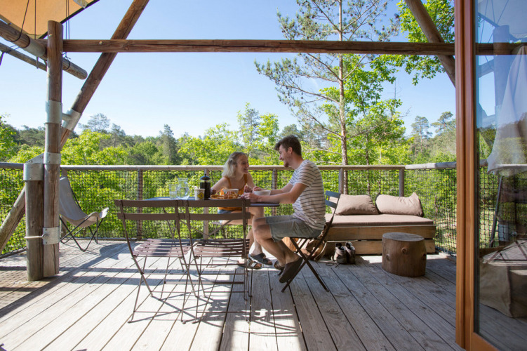 Pareja disfruta de una comida en la terraza de madera de una tienda Cahutte, rodeada de árboles y naturaleza.
