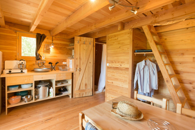 Acogedor interior de la tienda safari Cahutte, con paredes de madera, cocina sencilla y una escalera.