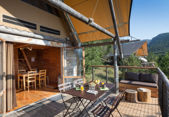 Terrasse extérieure d'une tente safari Cahutte avec coin repas, canapé et vue sur les collines boisées.