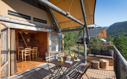 Buitenterras bij een Cahutte-safari tent met eethoek, bank en uitzicht op beboste heuvels.