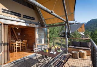 Buitenterras bij een Cahutte-safari tent met eethoek, bank en uitzicht op beboste heuvels.