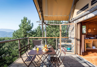 Zona pranzo esterna su terrazza in legno di una tenda Cahutte con vista su foresta e montagne.