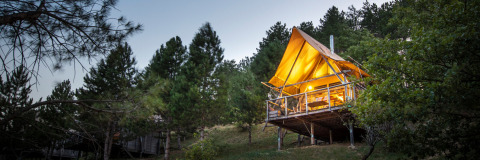 Illuminated safari tent called Cahutte in a forest clearing, elevated on stilts, surrounded by pine trees.