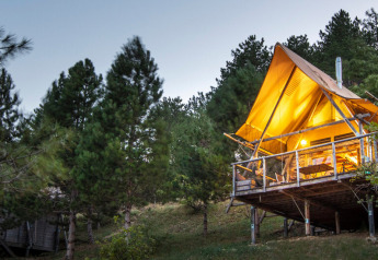 Illuminated safari tent called Cahutte in a forest clearing, elevated on stilts, surrounded by pine trees.