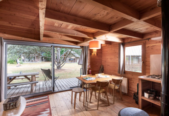 Intérieur de cabane chaleureux avec table à manger en bois, grandes fenêtres sur terrasse et jardin extérieur.