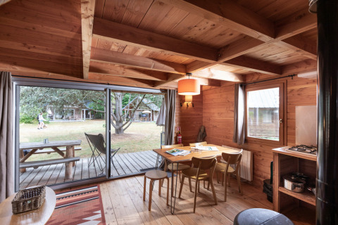Cozy cabin interior with a wooden dining table, large windows facing a deck and grassy yard area outside.