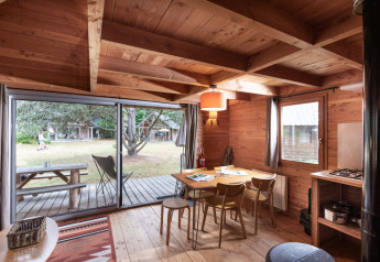 Intérieur de cabane chaleureux avec table à manger en bois, grandes fenêtres sur terrasse et jardin extérieur.