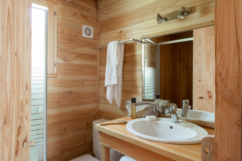 Baño de cabaña acogedor con paredes de madera, lavabo blanco, espejo, toalla y ventana luminosa.