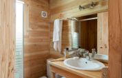 Baño de cabaña acogedor con paredes de madera, lavabo blanco, espejo, toalla y ventana luminosa.