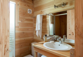 Baño de cabaña acogedor con paredes de madera, lavabo blanco, espejo, toalla y ventana luminosa.