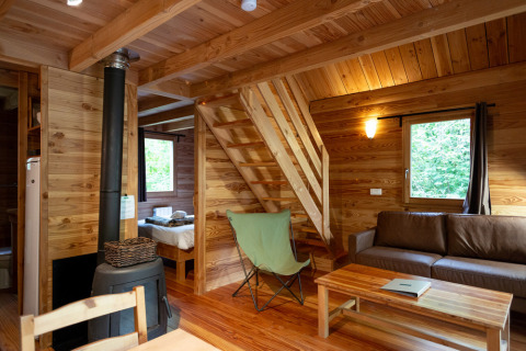 Intérieur chaleureux de cabane en bois avec poêle, canapé, escalier et fenêtre donnant sur la verdure.