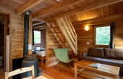 Acogedora cabaña de madera con estufa, sofá marrón, mesa, escalera y ventana hacia la naturaleza.
