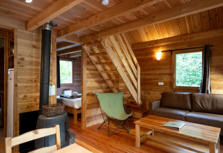 Acogedora cabaña de madera con estufa, sofá marrón, mesa, escalera y ventana hacia la naturaleza.