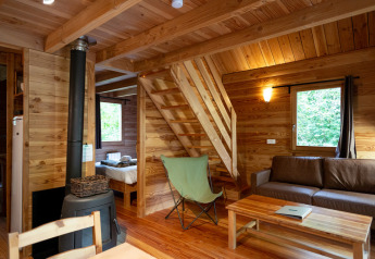 Interno di una baita in legno con stufa, divano marrone, tavolino, scala e finestra sulla natura.