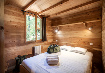 Chambre chaleureuse dans une cabane en bois avec lit double, fenêtre ouverte et sacs sur le lit.