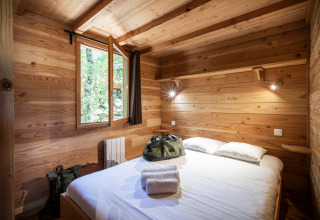 Acogedor dormitorio en una cabaña de madera con cama doble, ventana abierta y bolsas sobre la cama.