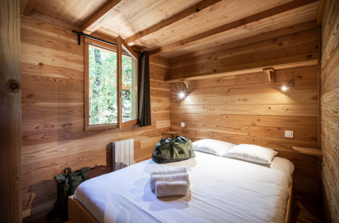 Cozy bedroom inside a wooden cabin with a double bed, open window, and bags on the bed in a forest setting.