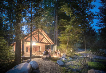 Cabane en bois chaleureuse dans la forêt au crépuscule, des personnes réunies autour d'un feu de camp.