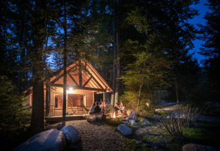 A cozy wooden cabin in the forest at dusk, with people gathered around a campfire in front.