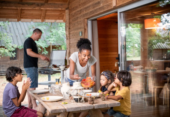 Une famille se retrouve sur la terrasse d’un chalet : un homme cuisine au barbecue, une femme sert les enfants.