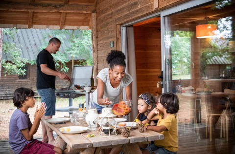 Op een terras aan een blokhut bereidt een man eten op de barbecue, terwijl een vrouw kinderen bedient aan tafel.