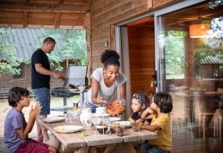 En familie hygger sig på terrassen ved en træhytte; en mand griller, mens børn og en kvinde er ved bordet.