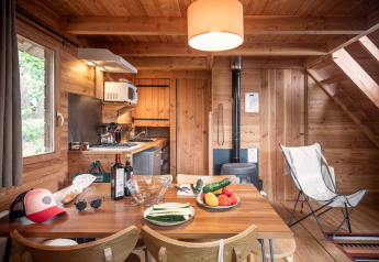 Intérieur de cabane en bois, table à manger avec légumes, vin, cuisine équipée et poêle à bois derrière.