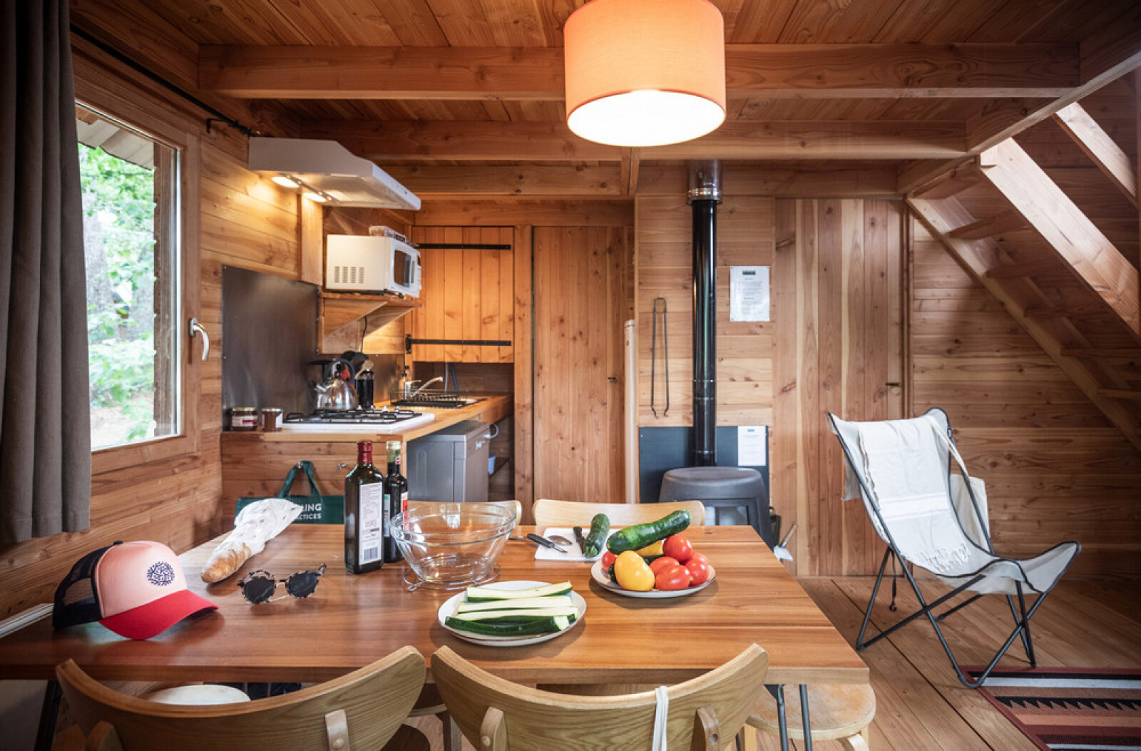 Knusse houten blokhut met eettafel vol groenten, wijn, keuken op de achtergrond en houtkachel zichtbaar.