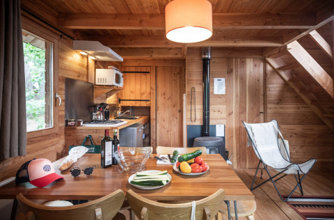 Hyggelig hytte med træinteriør, spisebord med grøntsager, vin, køkken og brændeovn i baggrunden.