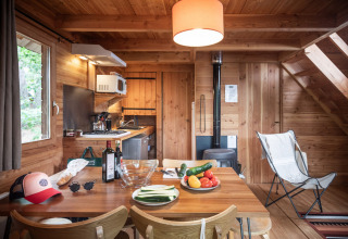 Hyggelig hytte med træinteriør, spisebord med grøntsager, vin, køkken og brændeovn i baggrunden.