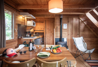 Hyggelig hytte med træinteriør, spisebord med grøntsager, vin, køkken og brændeovn i baggrunden.