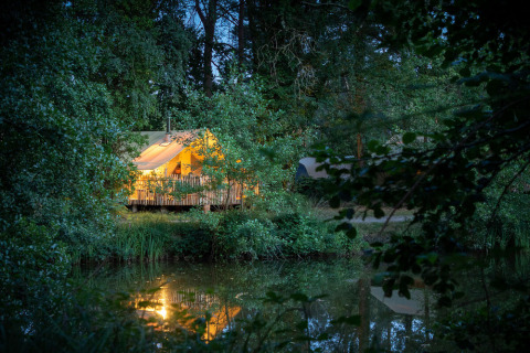 Tente safari éclairée au bord d’un lac forestier au Village Huttopia Lac de Rillé, France, en soirée.