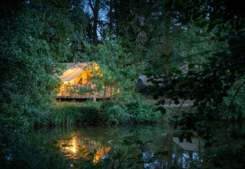 Tenda safari illuminata vicino al lago nel bosco al Village Huttopia Lac de Rillé, Francia, di sera.