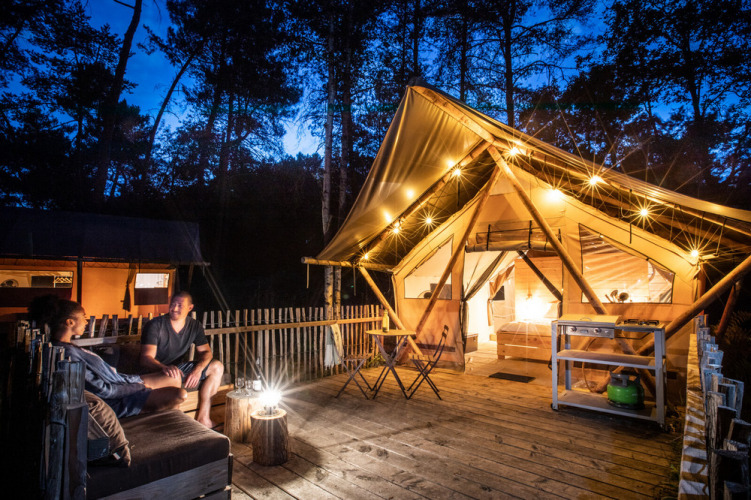 Two people relaxing outside a lit-up safari tent at Village Huttopia Lac de Rillé in France at night.