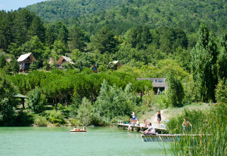 People relaxing by a lake with forest cabins at Village Huttopia Dieulefit holiday park in Auvergne-Rhône-Alpes, France.