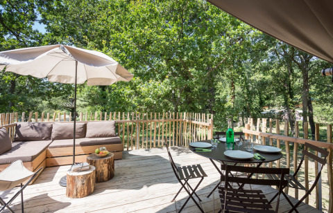 Terraza de madera en Huttopia Dieulefit con sofá, sombrilla, mesa para comer y vistas a un bosque verde.