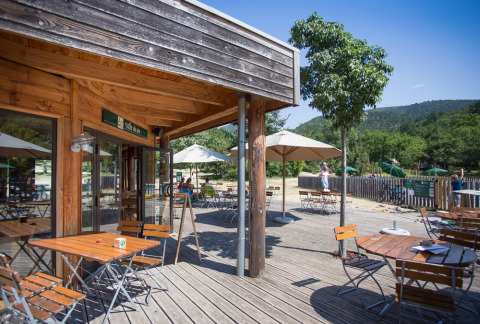 Terraza exterior en Village Huttopia Dieulefit, parque vacacional en Auvergne-Rhône-Alpes, con mesas y sombrillas de madera.