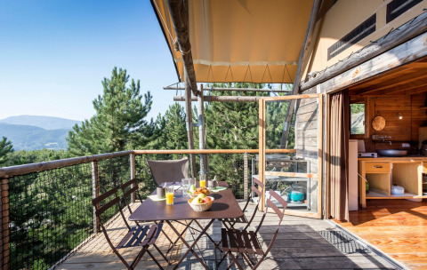 Wooden terrace with table and chairs overlooking pine trees at Village Huttopia Dieulefit, Auvergne-Rhône-Alpes, France.