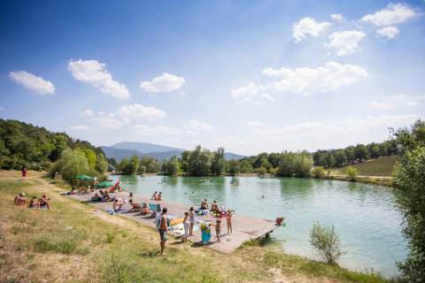 Holiday park by a lake at Village Huttopia Dieulefit in Auvergne-Rhône-Alpes, France, with people sunbathing.