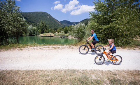 Padre e hija montan en bicicleta junto a un lago en Village Huttopia Dieulefit, rodeados de montañas y árboles.