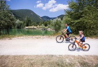 Padre e hija montan en bicicleta junto a un lago en Village Huttopia Dieulefit, rodeados de montañas y árboles.