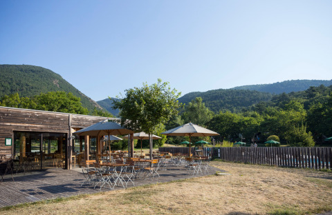 Zona exterior con mesas, sillas y sombrillas en Village Huttopia Dieulefit, Auvergne-Rhône-Alpes, Francia, entre montañas verdes.